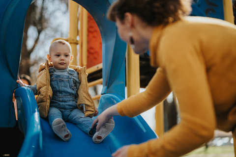 Child sliding down a playground slide with an mom nearby