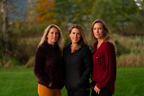 Three women standing together in a grassy field with trees in the background